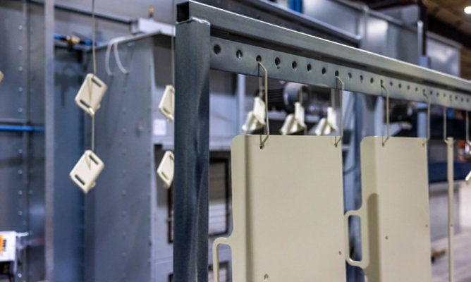 Metal components hang from an overhead conveyor rack inside an industrial powder-coating or finishing facility. 