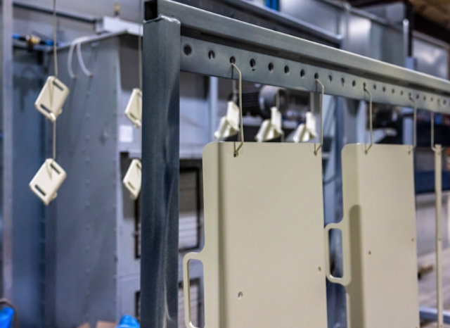 Metal components hang from an overhead conveyor rack inside an industrial powder-coating or finishing facility. 