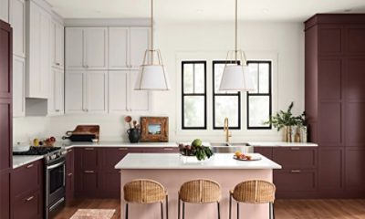 A large modern kitchen with white walls and cabinets in one section with maroon cabinets with gold handles in another. An island with a salmon colored base with 3 wicker bar stools.