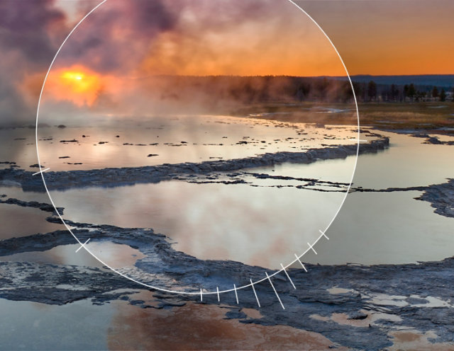 Photo of an erupting geyser surrounded by a pool of water with a sun setting in the background and an aperature dial focused on the geyser pool