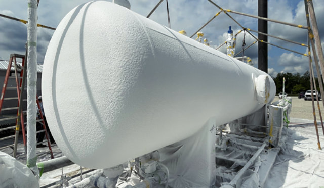 Large horizontal heater treater vessel on an outdoor industrial site, fully coated in white Heat‑Flex AEB, with surrounding piping, scaffolding, and protective sheeting visible.