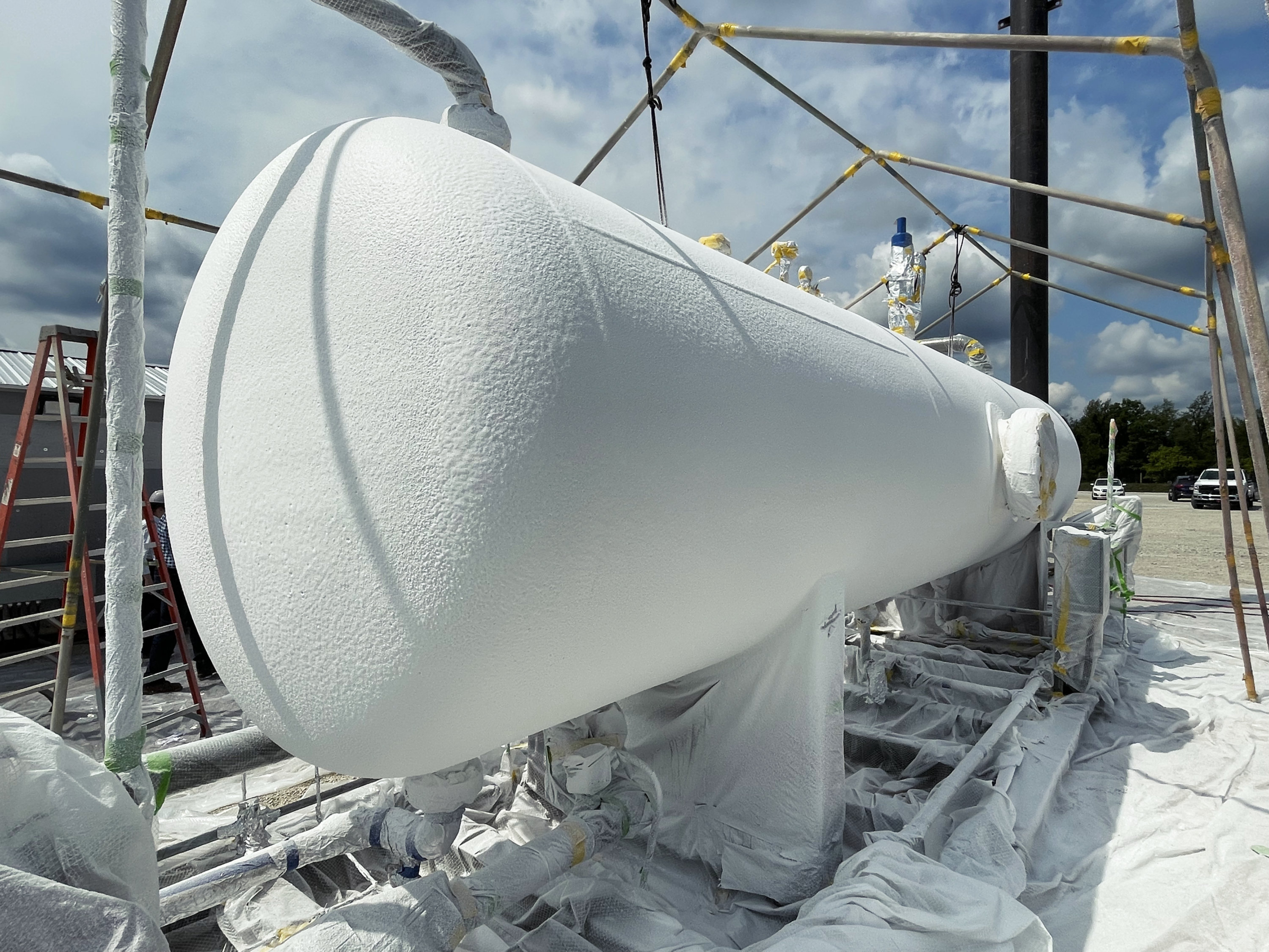 Large horizontal heater treater vessel on an outdoor industrial site, fully coated in white Heat‑Flex AEB, with surrounding piping, scaffolding, and protective sheeting visible.