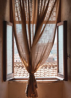 A sheer tan blush curtain tied in a knot hanging in the middle of a window looking out at a terracotta tiled roof.