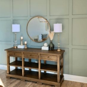 A console table stained a medium brown with lamps and décor on top against a wall painted a green-gray.
