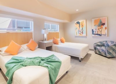 A Ronald McDonald House bedroom with two double beds, white bedding, orange pillows, a central nightstand with a lamp, abstract art on walls painted in Sherwin-Williams Pediment, and a blue plaid armchair.