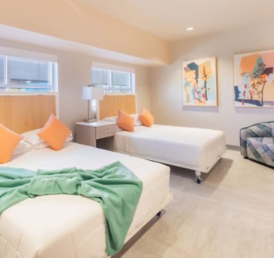 A Ronald McDonald House bedroom with two double beds, white bedding, orange pillows, a central nightstand with a lamp, abstract art on walls painted in Sherwin-Williams Pediment, and a blue plaid armchair.