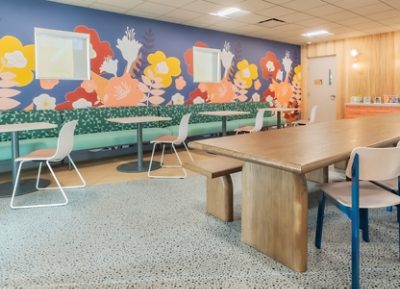 Ronald McDonald House dining area with a colorful floral mural, mint green banquettes, several small tables, and one large wooden table.