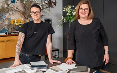 Christian Siriano and Sue Wadden, both dressed in black, standing at a table covered in paint samples in Siriano's home.