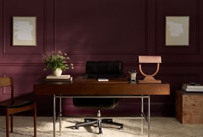 Office space with wooden desk, black leather chair, vase with flowers, modern lamp, and abstract art on walls painted in Sherwin-Williams Cordovan.