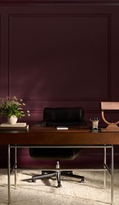 Office space with wooden desk, black leather chair, vase with flowers, modern lamp, and abstract art on walls painted in Sherwin-Williams Cordovan.