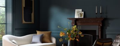 A stylish living room color-drenched in Sherwin-Williams Tarragon featuring a cream sofa, a carved wooden fireplace mantel, and a vase of orange flowers.