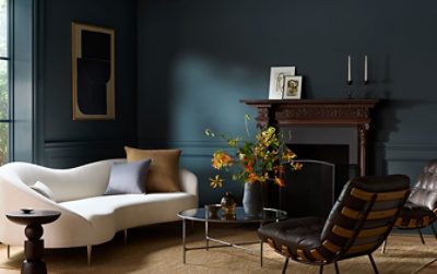 A stylish living room color-drenched in Sherwin-Williams Tarragon featuring a cream sofa, a carved wooden fireplace mantel, and a vase of orange flowers.