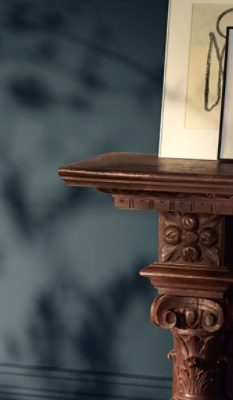 Ornately carved wooden fireplace mantel against a Tarragon-painted wall with plant shadows and framed line art visible on the mantel.