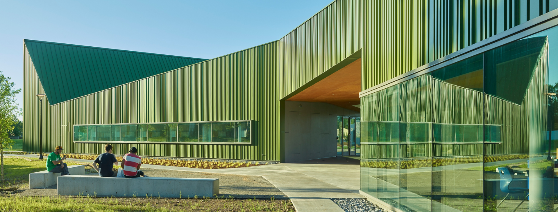 Exterior of Thaden school Reels and Wheels building in brilliant green metal with undulating roof in front of cloudless blue sky, students seated on cement benches in courtyard.