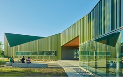 Exterior of Thaden school Reels and Wheels building in brilliant green metal with undulating roof in front of cloudless blue sky, students seated on cement benches in courtyard.