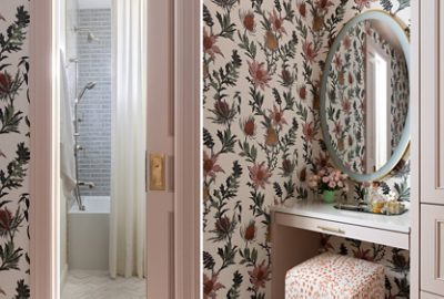 A stylish bathroom with trim, door, and built-in cabinets in Sherwin-Williams Abalone Shell, featuring a shower next to a floral wallpapered vanity area with a round mirror and a small vase of pink flowers.