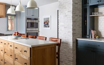 A modern kitchen featuring a light wood island with a bowl of fruit, brown leather chairs, and cabinetry in Sherwin-Williams Mineral Deposit alongside walls in Sherwin-Williams Drift of Mist and a bar in Sherwin-Williams Slate Tile.