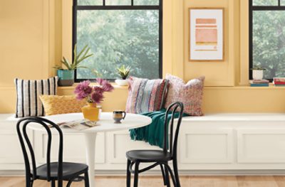 A breakfast area painted SW 7685 White Raisin, with a white circular table, 2 black chairs, white wooden wall bench with pillows and large window.