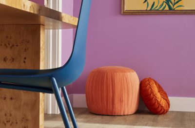 An orange pouf and darker orange velvet pillow on the floor in front of a bedroom wall painted Novel Lilac SW 6836.