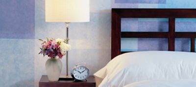 A closeup of a blue checkerboard painted bedroom wall with a clock on a nightstand, and bed with white pillow.