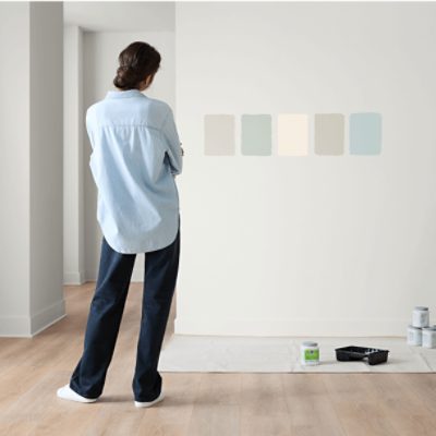 A person in a light blue shirt stands in front of a wall with five paint color samples. Painting supplies, including cans and a tray, are on the floor.