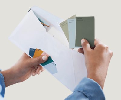 Hands holding a white envelope and pulling out several paint color chips in muted green and blue shades.