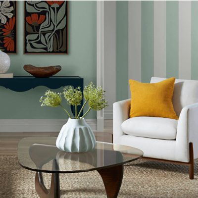 A modern living room with a light green and white striped wall. A white armchair with a yellow pillow sits beside a glass coffee table with wooden legs. On the table is a white vase with green flowers. In the background, a dark blue console table holds decorative items, with two framed floral artworks hanging above it.