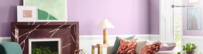 A living room with a purple Radiant Lilac SW 0074 wall, a red marble fireplace, green armchair, orange ottoman, gray sofa with patterned pillows, and a glass coffee table.