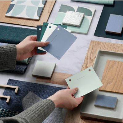 A person holds green and blue paint swatches over a background of fabric samples, tiles, and wooden surfaces.