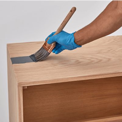 A gloved hand uses a paintbrush to apply gray paint to the top of a wooden cabinet or shelf.