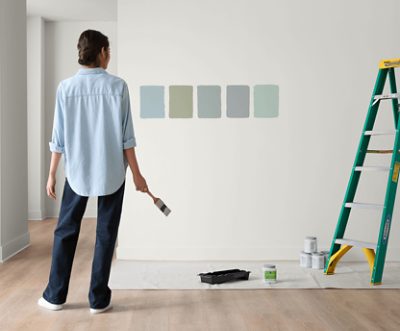 A person comparing wet paint samples applied to a wall with the used Color to Go containers sitting on the floor on a dropcloth.