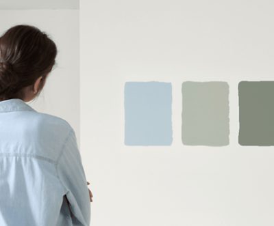 Person seen from behind looking at three painted color swatches on a wall in soft blue and green tones.