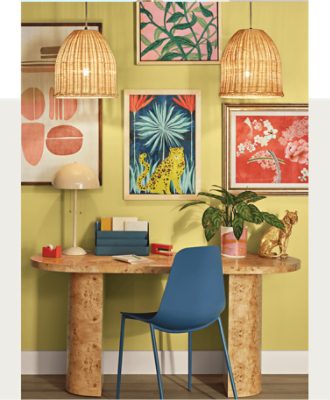 Home office with a wooden desk, blue chair, lamp, red stapler, papers, and a potted plant. Three colorful framed artworks and two wicker pendant lights hang above.
