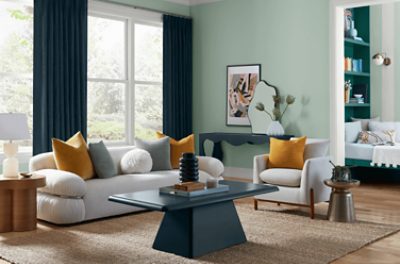Living room painted in SW 6456 Slow Green with neutral couch, throw pillows, side tables, and large windows.