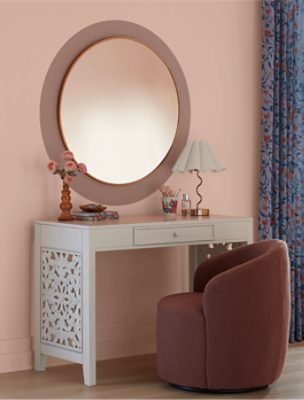 Wall painted in SW 6610 Koral Kicks, small round chair facing a white desk with a round mirror hung up.
