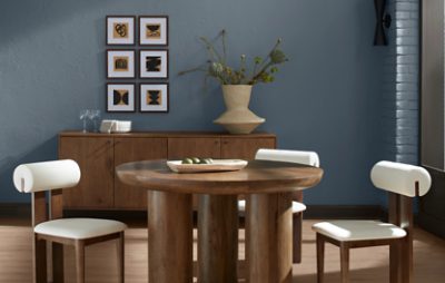 Round wooden table with chairs with white cushions, wooden credenza behind it with some artwork hung on a wall painted in SW 6249 Storm Cloud.