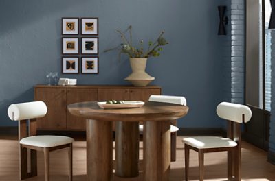 Round wooden table with chairs with white cushions, wooden credenza behind it with some artwork hung on a wall painted in SW 6249 Storm Cloud.