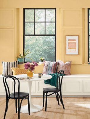 A breakfast area with a white circular table, 2 black chairs, white wooden wall bench with pillows and large window.