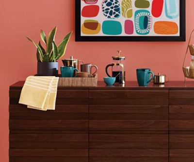 Dark wooden drawers with a plant and mugs on it under a frame photo.