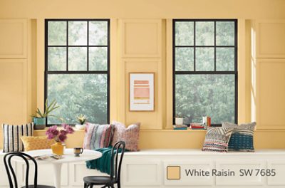 A breakfast area with a white circular table, 2 black chairs, white wooden wall bench with pillows and large window.