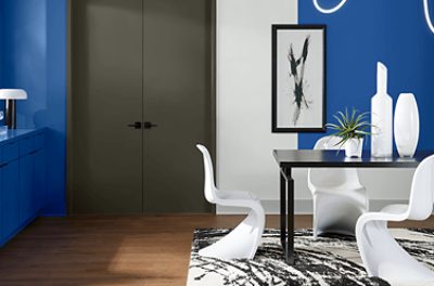 Wall painted in SW 6967 Frank Blue with a simple black frame, black table with white chairs and decor, area rug underneath, gray double doors to the left of the wall.