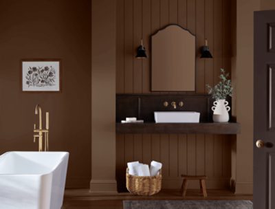 Modern bathroom with brown walls, white freestanding tub, dark wood vanity with gold fixtures, arched mirror, and decorative accents like plants and towels.