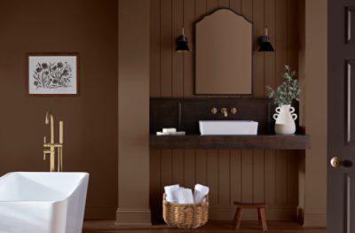 Modern bathroom with brown walls, white freestanding tub, dark wood vanity with gold fixtures, arched mirror, and decorative accents like plants and towels.