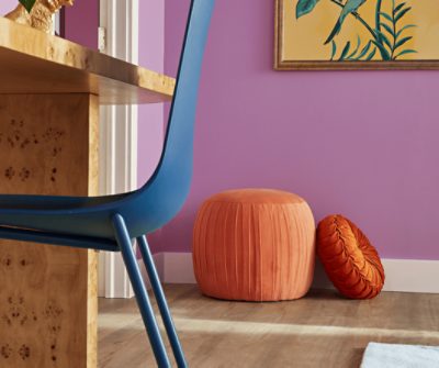 An orange pouf and darker orange velvet pillow on the floor in front of a bedroom wall painted Novel Lilac SW 6836.