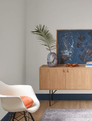 A white chair near a light wood sideboard with a large blue abstract print on top against walls painted Snowbound SW 7004.