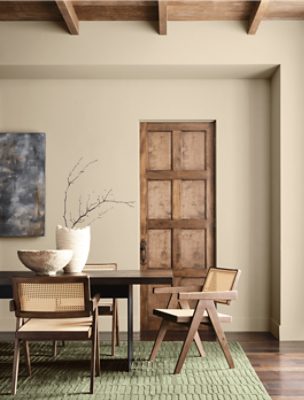 Interior with a dark wooden table, light wooden chairs, ceramic bowl, and vase with branches in front of a Universal Khaki wall and wooden door.