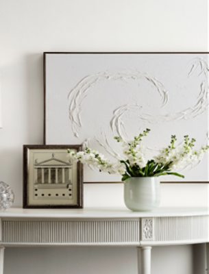 A white side table in front of a white wall with flowers and artwork on top.