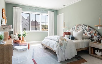 Soft green bedroom on a sunny day with lots of throw pillows and blankets on bed with oversized arch fabric-covered headboard, neutral accents and white curtains.