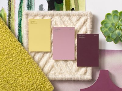 Yellow, pink, and berry colored paint chips laid out on a cream woven material, near other fabrics and a plant.