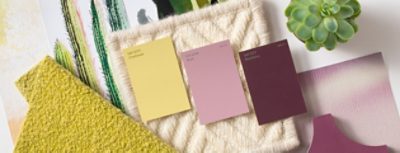 Yellow, pink, and berry colored paint chips laid out on a cream woven material, near other fabrics and a plant.